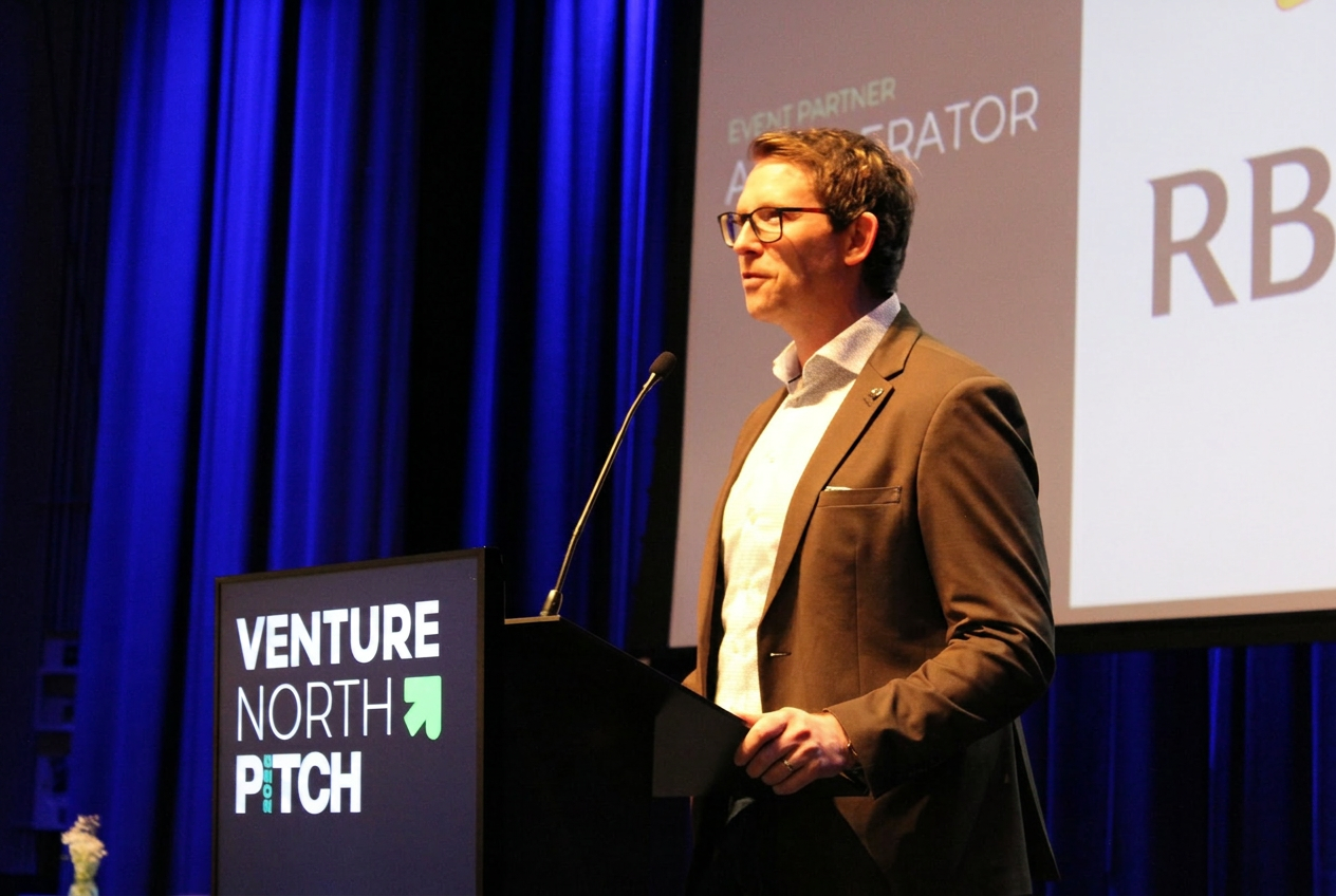 Speaker at the Venture North PITCH podium with Event Partner sponsor branding projected on stage.